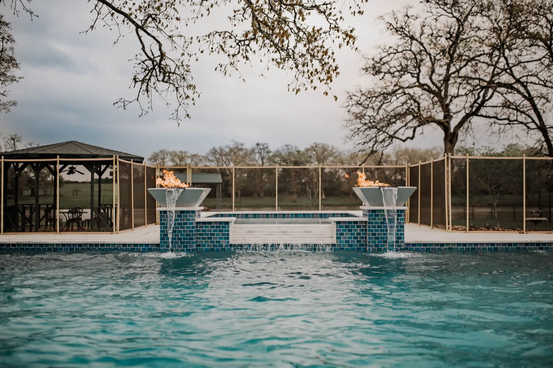 A luxurious pool features two flame-lit fountains, surrounded by a safety fence and a shaded seating area in a serene outdoor setting