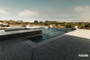 A modern swimming pool surrounded by a stone deck, patio chairs, and a serene natural landscape under a clear sky