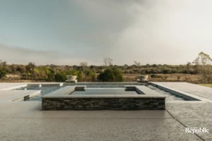 A serene outdoor pool area featuring a hot tub, stone accents, and wooden chairs, surrounded by lush landscaping under a soft sky